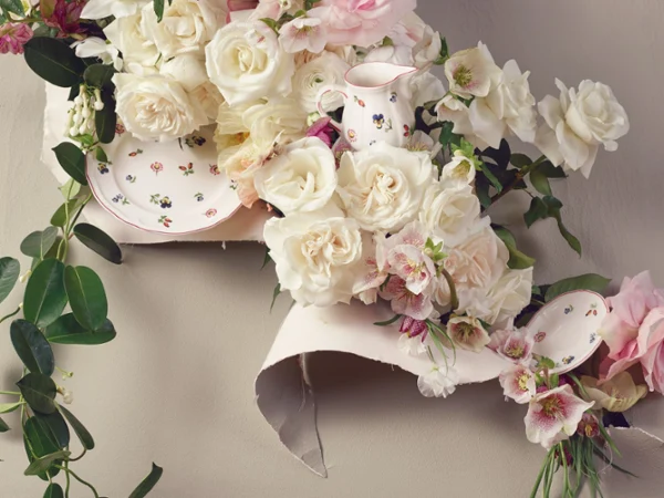 Petite Fleur in a floral arrangement Elegant floral arrangement featuring white and pink roses intertwined with green leaves, surrounded by a floral-patterned plate and pitcher. The scene conveys a romantic, vintage vibe.