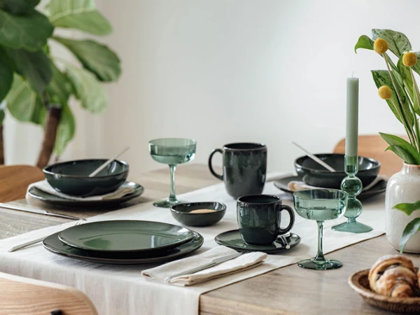 Una mesa de comedor con vajilla verde, cristalería, cubiertos, una vela y un jarrón con flores amarillas.