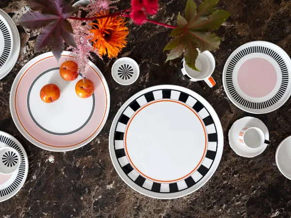 Memphis in allen Facetten Memphis Tischgedeck von Villeroy & Boch mit dekorativen Tellern, einem Becher, Tassen und Pflaumen auf einer mit leuchtenden Blumen geschmückten braunen Marmoroberfläche.