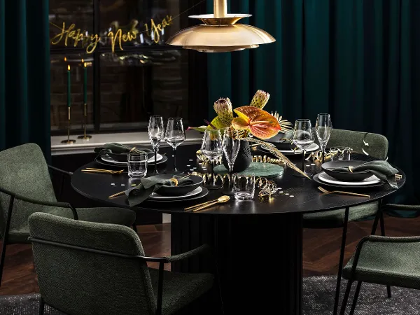 Celebrating New Year’s Eve with Villeroy & Boch Manufacture A round black dining table is elegantly set with green napkins, gold cutlery and a flower arrangement. Green chairs surround the table and a "Happy New Year" sign welcomes guests to the New Year's Eve party.