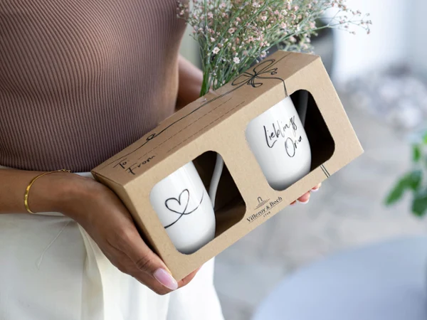 Dos tazas con la inscripción "Lieblingsoma" (abuela favorita) en una caja de regalo Una persona sostiene una caja con dos tazas Statement con la inscripción "Lieblingsoma" (abuela favorita) y un ramo de flores pequeñas.