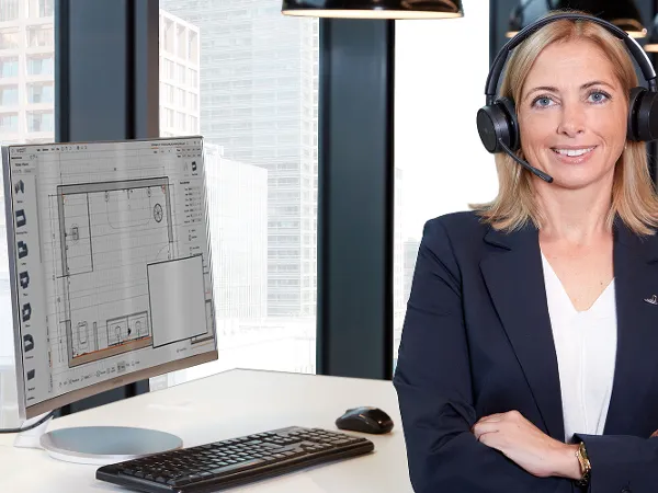 Employée de Villeroy & Boch pour le conseil en salle de bain Une employée est assise à son bureau face à la caméra. En arrière-plan, on peut voir la planification de la salle de bain sur l'écran