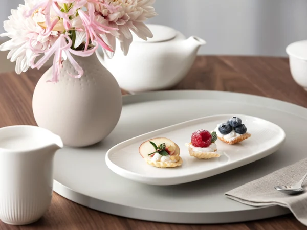 A white Afina plate with three small pastries and fruit sits on a round tray next to a flower vase, a teapot and a milk jug on a wooden table.
