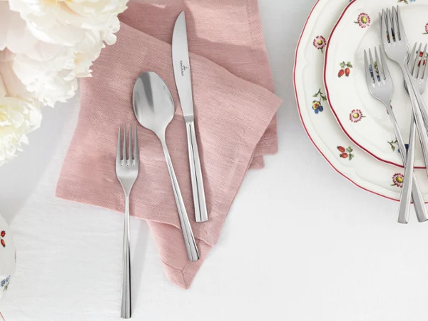 Piemont cutlery lies on a pink Lifestyle Accessories napkin. Piemont cutlery lies on a pink Lifestyle Accessories napkin next to stacked Petite Fleur plates on a white tablecloth.