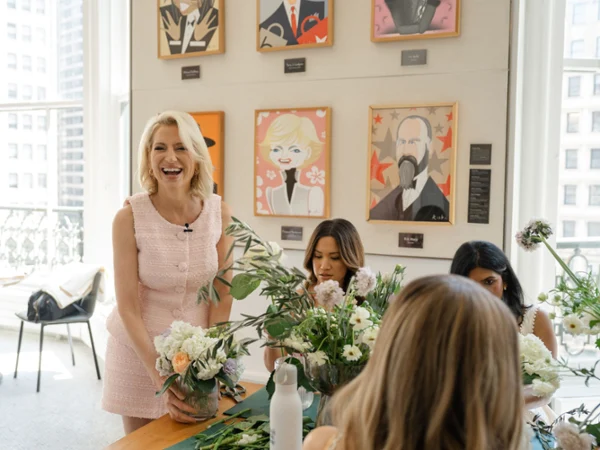 Dorinda Medley A woman, Dorinda Medley, in a pink dress smiles while arranging flowers on a table with three others. Colorful portraits hang on a wall, with large windows in the background.