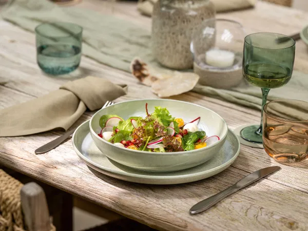 Un bol de la collection Perlemor de Villeroy & Boch avec une salade fraîche La collection Perlemor de Villeroy & Boch avec une salade fraîche composée de différents légumes dans un saladier sur une table en bois, complète avec des couverts, des verres et des serviettes beiges.