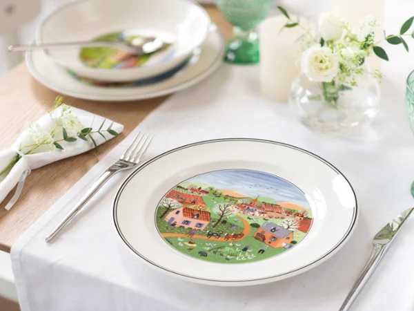 design-naif-dinner-placesetting-with-new-moon-flatware Elegant table set with white dishes, featuring a colorful countryside scene with houses and trees. Accented by green glassware and white floral decor.