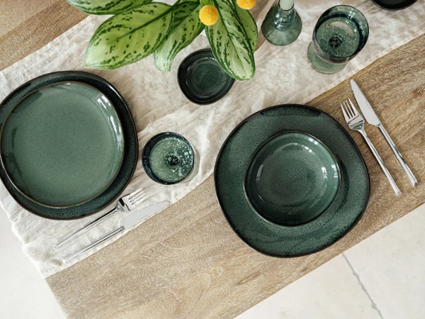 Stylishly set wooden table with "Lave Vert" ceramic tableware, silver "like cutlery" from like. by Villeroy & Boch, and matching Like glasses on a cream-coloured fabric napkin. A green plant with yellow accents complements the elegant arrangement.