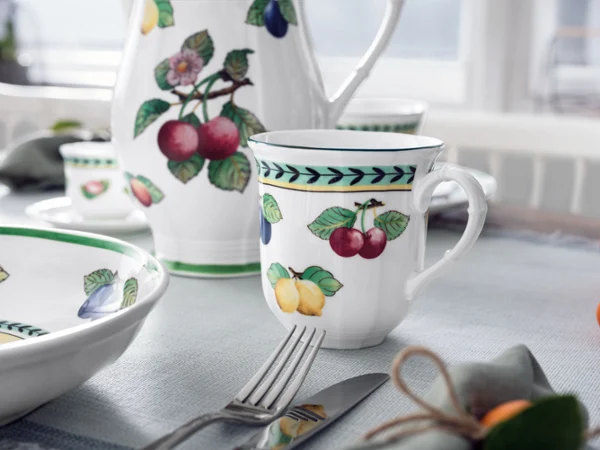 French Garden Mug on table with pitcher and bowl A fruit-patterned porcelain mug from the French Garden collection displayed alongside matching dinnerware and a coffee pot on a bright table setting.
