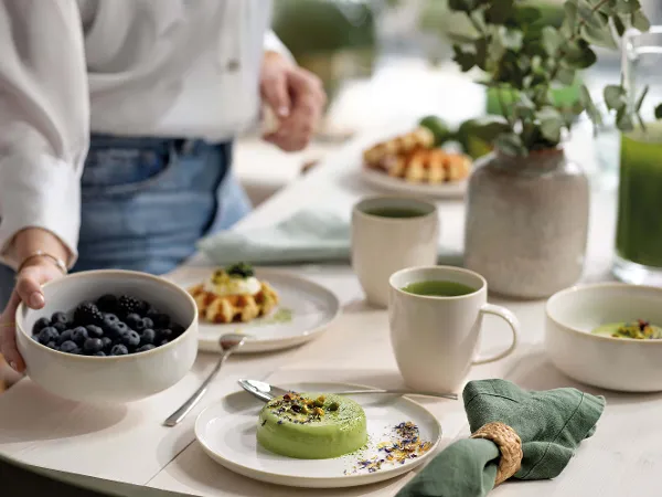 Des assiettes pour le petit-déjeuner Crafted Cotton avec de la nourriture Une personne prépare une table pour le petit-déjeuner avec différents plats servis dans des assiettes pour le petit-déjeuner Crafted Cotton et des tasses à thé.
