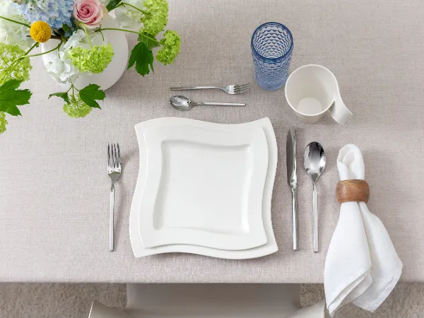 Modern top view of a single table setting featuring NewWave porcelain Clean, modern top view of a single table setting featuring NewWave porcelain plates and cup, Boston Blue long drink glass, and matching NewWave cutlery. Neutral tones and minimalist aesthetic create a contemporary dining impression.