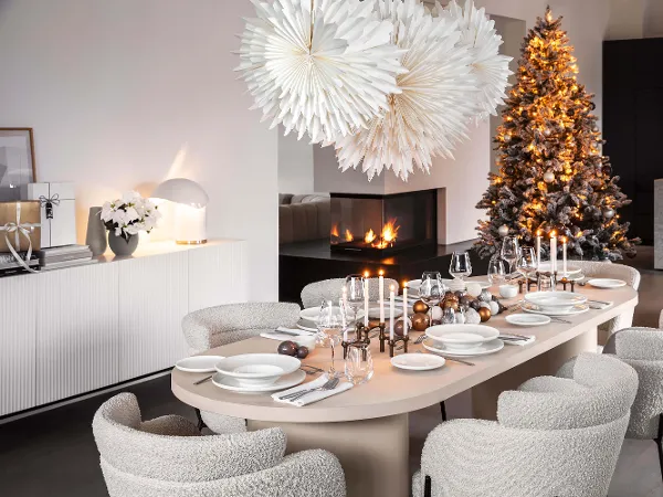 De la vaisselle Afina dans une ambiance de Noël moderne Une salle à manger moderne est couverte d’assiettes Afina blanches et de bougies pour un repas. Il y a également deux grandes étoiles en papier pour la décoration, un sapin de Noël décoré et une cheminée intégrée.