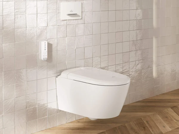WC douche Supreo sur un mur carrelé blanc Un WC douche moderne et blanc est fixé à un mur carrelé au-dessus d’un sol en bois à motif chevrons.