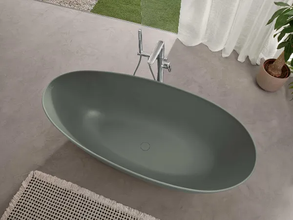 Une baignoire verte Antao et une robinetterie sur pied en chrome Une baignoire ovale verte de la collection Antao de Villeroy & Boch se trouve sur un sol carrelé, avec un palmier à côté.