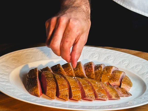 Close-up of a chef arranging sliced roasted duck on a white platter, with golden-brown skin, pink centers, and dipping sauces below.