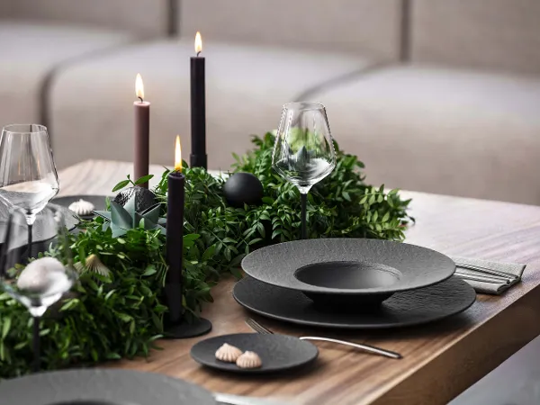Des assiettes noires Manufacture sur une table de Noël au design moderne Une table de salle à manger avec des assiettes noires Manufacture, des couverts de table, des verres à vin et des bougies sombres. Des plantes vertes sont utilisées comme décoration de table et créent une atmosphère raffinée.