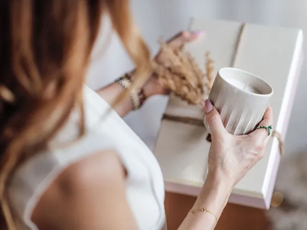Une personne tient une bougie parfumée Perlemor dans sa main gauche. Une personne tient une bougie parfumée Perlemor dans sa main gauche et un cadeau emballé, décoré de plantes séchées.