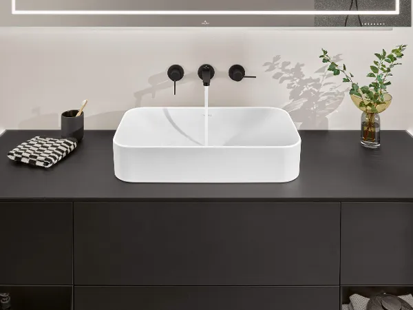 Un lavabo Finion blanc sur un meuble noir Un lavabo de salle de bains moderne au design carré sur un plan de toilette noir, un robinet chromé et une plante décorative