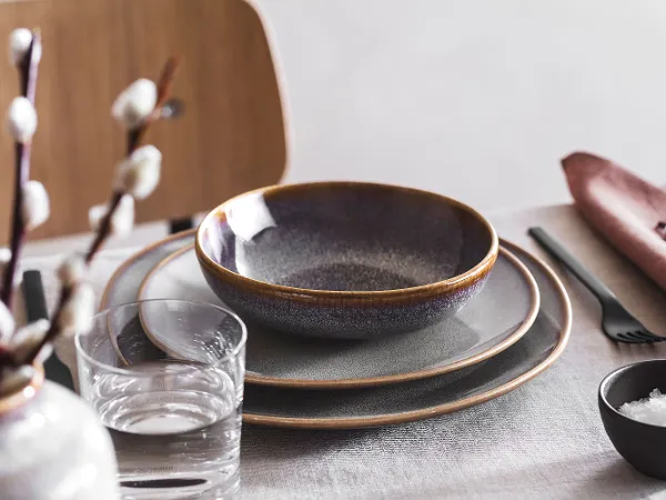 Elegant table setting with stacked Lave plates, a bowl, flatware, and a glass of water next to a flower arrangement.