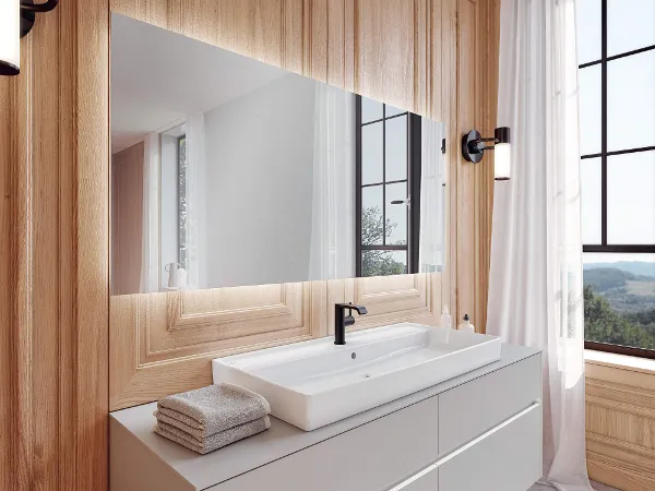 Un miroir « More to See Lite » sur mur en bois au-dessus d’un grand meuble blanc Une salle de bains moderne avec un grand lavabo blanc posé sur un meuble en bois, flanquée de hautes fenêtres offrant une vue pittoresque. Des serviettes moelleuses sont parfaitement empilées sur le côté.
