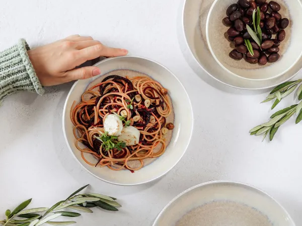 Hand reaches for Perlemor pasta bowl with pasta, surrounded by olives in small bowls on white table