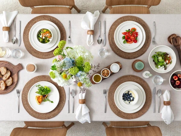 Top view of a beautifully set dining table with salads, fruit, bread, water glasses, and flowers in the centre on a beige tablecloth.