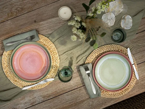 Deux couverts dressés avec Perlemor sur une table en bois Deux couverts dressés avec Perlemor sur une table en bois avec des sets de table ronds tissés, des couverts de table, une bougie, des fleurs et des plantes vertes.