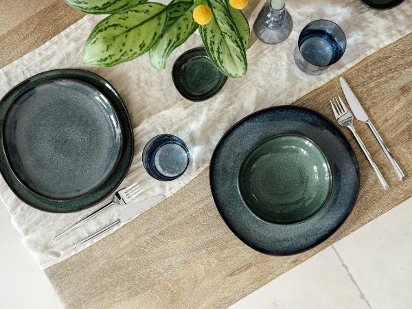 Deux couverts Lave avec des assiettes vertes, des couverts et des verres bleus décorent une table en bois Deux couverts de Villeroy & Boch Lave avec des assiettes vertes, des couverts et des verres bleus décorent une table en bois avec un chemin de table blanc. Au centre, une plante à feuilles vertes est placée dans un vase.
