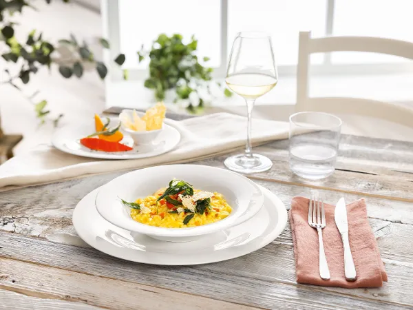 Die New Cottage-Kollektion von Villeroy & Boch auf einem rustikalen Holztisch. Eine Schüssel cremiges Kräuterrisotto steht neben einer kleinen Vorspeise, einem Glas Weißwein und Besteck auf der New Cottage-Kollektion von Villeroy & Boch auf einem rustikalen Holztisch.