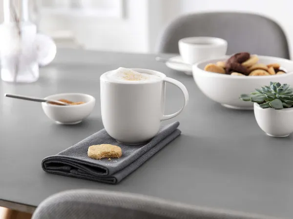 Un mug NewMoon avec une coupe et de la pâtisserie Une tasse à cappuccino sur une serviette grise avec un biscuit, des bols avec des snacks et une succulente sur une table de salle à manger moderne.