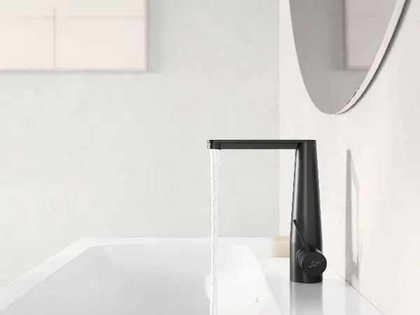 Black Conum washbasin tap with running water A modern black tap from which water is flowing into a white washbasin. A partially visible mirror on the right-hand side reflects the scene.