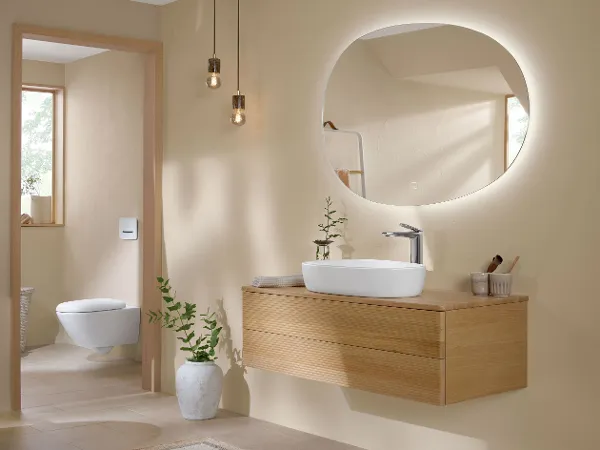 Antao Badezimmer mit Oak Holz-Badmöbeln und weißer Keramik in typischer Tropfenform Ein modernes helles Badezimmer mit einem runden, hinterleuchteten Spiegel über einem schwebenden Holzwaschtisch mit weißem Becken. Von der Decke hängen zwei Pendelleuchten. Auf dem Boden neben dem schwebenden Waschtisch steht eine Topfpflanze.