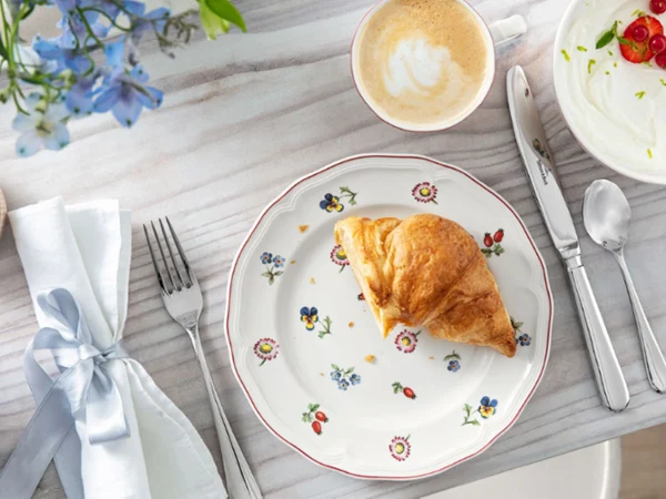 petite fluer bread plate A croissant on a flower plate with coffee, berries, yogurt, cutlery and a napkin with a bow.