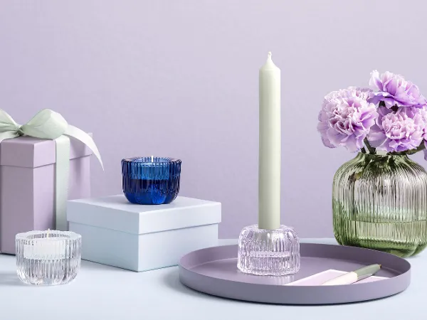 Drei Villeroy & Boch Fleur-Kerzenhalter aus Glas und eine Fleur-Vert Vase Die Villeroy & Boch Fleur Vert Vase mit lila Blumen wird auf einer Fläche neben Geschenkboxen und Fleur Kerzenhaltern präsentiert.