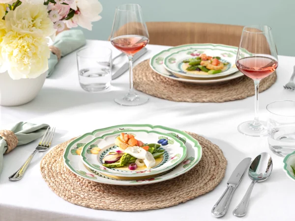 French Garden by Villeroy & Boch, modernly interpreted A set French Garden table for two people with pasta dishes, rosé glasses, water glasses, and a salad on woven placemats.