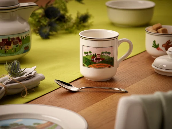 design-naif-mug-on-wooden-table A mug with a countryside illustration on a wooden table. Surrounded by a teapot, spoon, and napkin on a green tablecloth. Cozy and inviting setting.