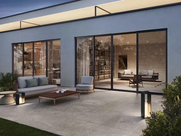 Moderne Terrasse mit Lucca Outdoor Fliesen Moderne Terrasse mit beigen Steinfliesen, grauen Gartenmöbeln und beleuchteten Laternen, neben großen Glasschiebetüren, die im Inneren den Blick auf einen zeitgenössischen Wohn- und Essbereich freigeben.