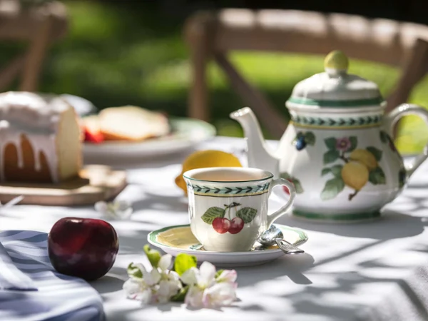 French Garden coffee cup on a white tablecloth Outdoor table setting with tea and snacks, with of a patterned teapot, a teacup, cake and fruit in natural daylight.