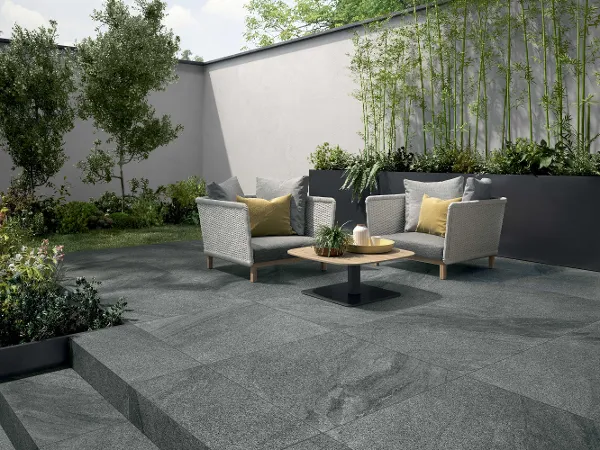 Außenterrasse mit Mont Blanc Garden Fliesen Außenterrasse mit zwei hellen Stühlen, einem runden Holztisch und grauen Steinfliesen. Grüne Pflanzen und Sträucher in Pflanzkübeln und entlang der Wand sorgen für Dekoration.