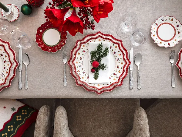 Festive Toy's Delight crockery setting Festive table setting with decorative Toy's Delight plates with red and white patterns, silver cutlery and a centrepiece of red flowers and green plants on a beige tablecloth.