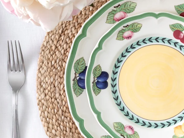 French Garden place setting close-up A white ceramic plate with a green scalloped edge and illustrations of blueberries, cherries, and peaches sits on a round woven tan placemat. A silver fork lies on a white tablecloth to the left, with white and pink flower petals in the upper left corner.