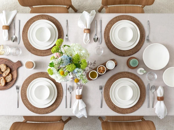 Artesano – A collection in pure white An elegantly set dining table with woven placemats, white plates, napkin rings, and flowers as table decorations. Bread on a wooden tray and small bowls are also on the table.
