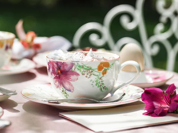 A Villeroy & Boch Mariefleur Basic teacup on a saucer, filled with coffee and whipped cream, on a table beside a flower. There is a white wrought-iron chair in the background.