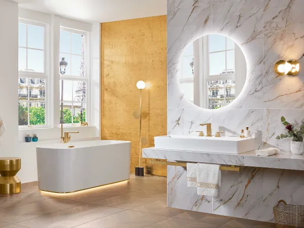 Une baignoire en îlot Mettlach de Villeroy & Boch, un grand miroir rond, un plan double et touches de doré Une salle de bains moderne avec une baignoire en îlot Mettlach de Villeroy & Boch, des murs en marbre, un grand miroir rond, un plan double et des touches de doré.