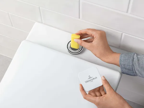 Vullen van de ViFresh-voorziening van een toilet Een persoon gooit een gele toiletsteen in de stortbak van een toilet. Op de achtergrond zijn witte betegelde muren te zien.