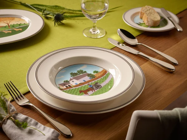 A table setting with a bowl depicting a farm scene, surrounded by cutlery, a napkin and a bread plate.