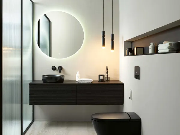 Schwarze Antao Toilette und Waschbecken von Villeroy & Boch Modernes Badezimmer mit rundem, von hinten beleuchtetem Spiegel, schwarzem, schwebendem Waschtisch mit Aufsatzwaschbecken, schwarzer Antao Toilette von Villeroy & Boch und minimalistischem Dekor. Von der Decke hängen zwei Pendelleuchten. Glastrennwand links.
