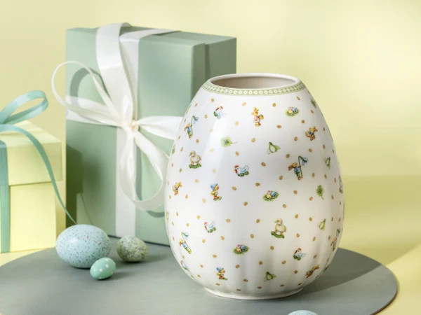 Un vase blanc Easter Delight avec des motifs floraux Un vase blanc Easter Delight avec des motifs floraux est exposé à côté de deux boîtes-cadeaux aux couleurs pastel et d'œufs mouchetés.