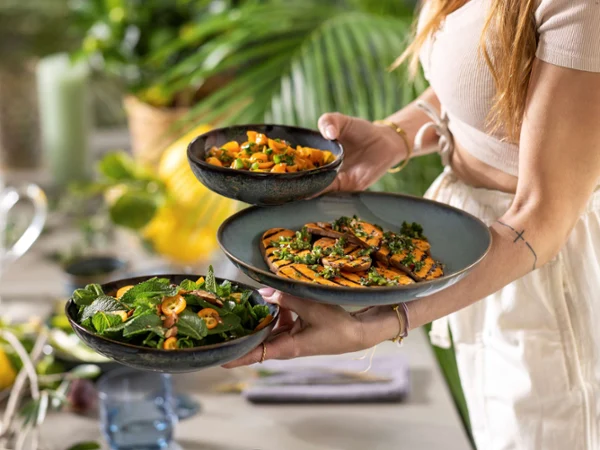 Serving Colorful Fresh Dishes A person holding three bowls of colorful dishes, including a salad with mint, roasted sweet potatoes, and a vegetable medley, in a lush setting.