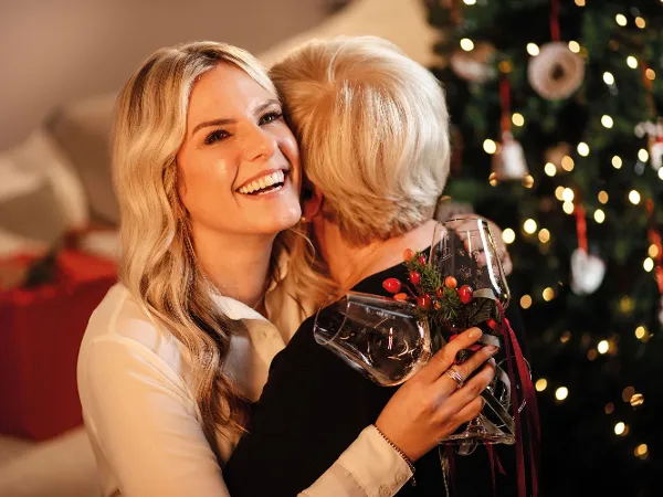 Two women embrace, one holding a Toy’s Delight wine glass. Two women embrace in front of a decorated Christmas tree. The woman looking into the camera is holding a Toy’s Delight wine glass.